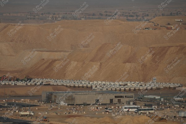A large construction site featuring heavy machinery and equipment, such as trucks and excavators, situated in front of large sandy hills. There are several metal structures and buildings under construction, possibly part of a major development project. Residential homes and buildings are visible in the background.