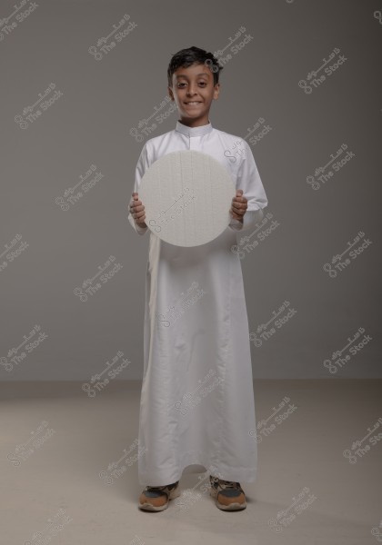 A child wearing a long white thobe, holding a large white circle in front of them, standing in an indoor setting with soft lighting, smiling gently.