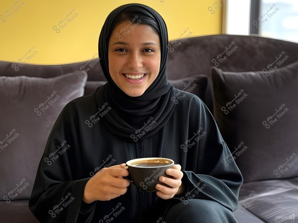 A portrait of a young woman wearing a black abaya and hijab, sitting on a dark-colored sofa with a yellow wall background. The woman is smiling and holding a cup of coffee in her hands. The setting appears to be cozy and naturally lit.