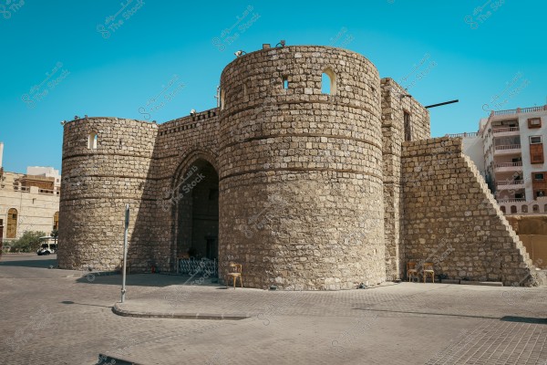 Image showing the historic Mecca Gate in Jeddah, Saudi Arabia. The gate is constructed from stone, featuring round towers on either side and a large arched entrance. Some chairs are placed nearby. Modern buildings are visible in the background under a clear blue sky.
