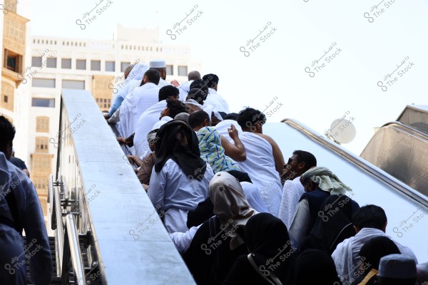 The image shows a large group of people ascending a staircase in one direction, most of them wearing white Ihram garments. Some women are seen wearing black abayas and veils, and there is a variety of other clothing suggesting visitors from diverse backgrounds and cultures. The image is likely taken in Mecca during the Hajj or Umrah season, with a modern building visible in the background.