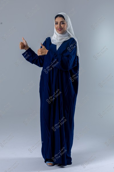 Image of a woman wearing a long blue abaya and a white hijab, standing against a neutral white background. The woman is smiling and giving a thumbs-up gesture with both hands. The attire appears to have a traditional Saudi or Gulf style.