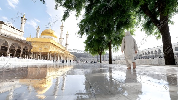 The image depicts the Grand Mosque in Mecca, featuring the golden dome and minarets, with green trees casting shade over the outer courtyard. A person wearing traditional white attire known as a thobe and bisht is walking barefoot on the shiny marble floor of the courtyard. Natural light reflects on the surface, with a gathering of people in the background.