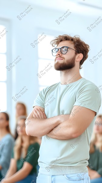 Image of a man wearing glasses and a light-colored shirt, standing with arms crossed, smiling confidently in a brightly lit room. In the blurred background, there are people sitting, dressed casually.