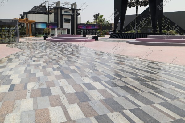 صورة تُظهر ساحة مفتوحة ذات تصميم أرضيات مبلطة بألوان متنوعة من الرمادي والبني. في الخلفية يوجد مبنى حديث مع واجهات زجاجية, وعلى الجانب الأيسر تظهر واجهة زجاجية أخرى بنمط زخرفي. توجد أيضًا منصات دائرية باللون الوردي الفاتح حول الساحة, بالإضافة إلى أشجار نخيل في الخلفية.