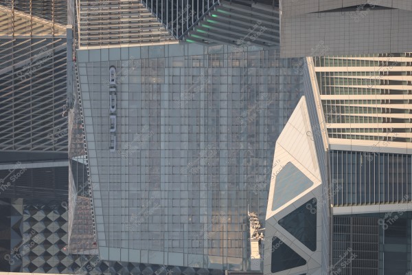 Image of a modern building with a glass facade reflecting the surrounding skyline. The facade features a complex design with slanted lines and geometric shapes, including large triangles. The building is surrounded by other architectural structures, indicating its placement in a developed urban area.