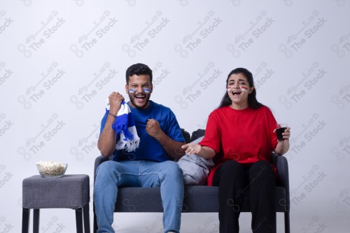 Portrait of a Saudi young man and woman watching a match and cheering their favorite teams cheerfully while having a snack on a blue background, the World Cup.