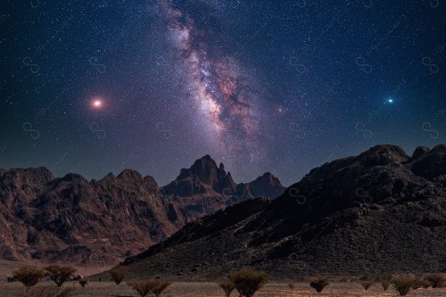 A series of rocky mountains and bushes in a desert whose sky is decorated with stars and planets.