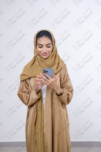 Portrait of a Saudi girl wearing an abaya on a white background holding a phone and making gestures with her hands while smiling, souvenir photos, documenting a happy moment.