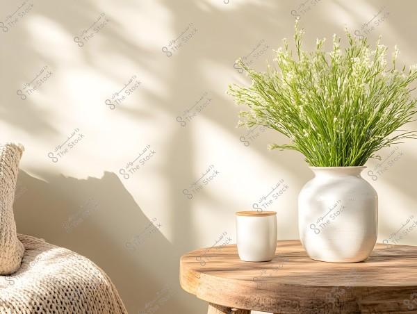 The image shows a large white vase filled with small green plants placed on a round wooden table. Next to the vase is a white cup with a wooden lid. In the background, there is a part of a sofa covered with a light beige knitted blanket, with sunlight casting soft shadows on the wall.