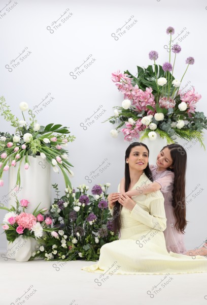 A woman in a long yellow dress sits on the ground while a young girl gently wraps her arms around her shoulders. Both are smiling softly. The background features beautiful floral arrangements with various colors such as white, pink, and purple. The scene conveys an atmosphere of tenderness and calm.