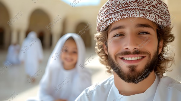 A portrait of a smiling man wearing a patterned brown headscarf and a white garment. In the background, there is a young boy also dressed in white, with several stone arches, suggesting a traditional architectural setting. The image conveys a cultural atmosphere typical of Oman or the Gulf region.