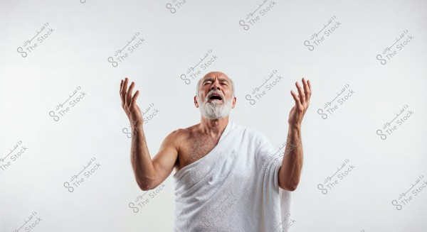 An image of an elderly man with a white beard, wearing a white wrap that covers his right shoulder, raising his hands with an expressive look on his face. The background is plain white, emphasizing the man\'s appearance. The wrap resembles the Ihram clothing used during Hajj.