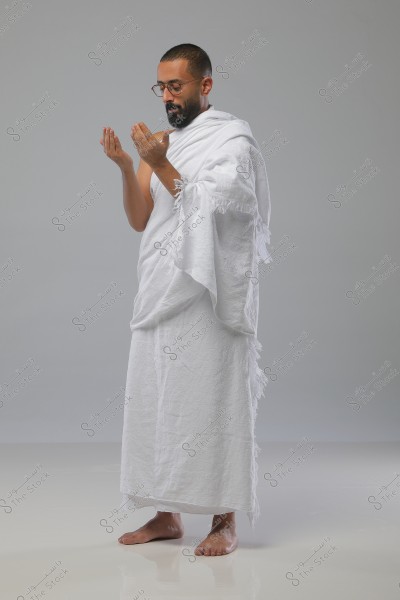 A portrait of a man wearing the white Ihram attire, possibly preparing for Umrah or Hajj. The man is standing and raising his hands in supplication. The background is neutral and simple, conveying a calm and spiritual atmosphere.