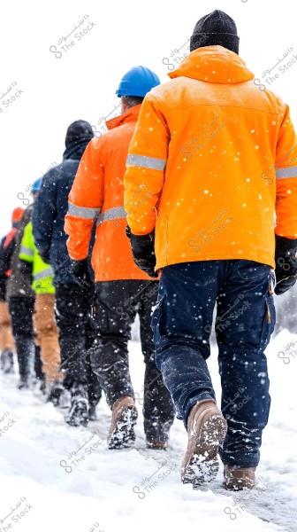 The image shows a group of workers walking in a line on snow. They are wearing orange and dark yellow jackets with reflective strips, and some are wearing blue helmets and warm hats. The atmosphere is cold with snowfall covering the boots and ground.