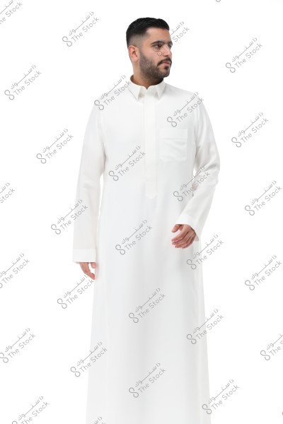 An image of a man wearing a traditional white thobe similar to a Saudi thobe. He stands against a white background, looking to the right. The thobe features a traditional collar and a pocket on the left side of the chest.