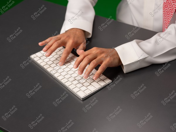 شخص يرتدي ثوبًا أبيض وكوفية حمراء يستخدم لوحة مفاتيح على مكتب.