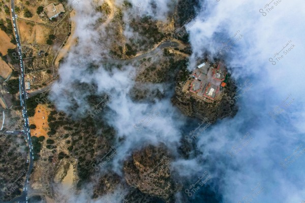 The aerial image shows a mountainous area covered with thick fog. A large building with red roofs is visible on the edge of a cliff on the right. Roads wind through the area filled with trees and vegetation. On the left, cars are lined up along a narrow road, and small houses are scattered throughout.
