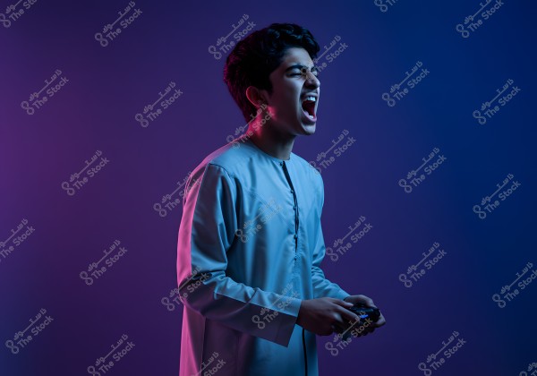 Portrait of a young man wearing a light-colored traditional thobe, appearing enthusiastic while holding a video game controller. The surrounding lighting is colored in shades of blue and purple, adding a dynamic atmosphere.