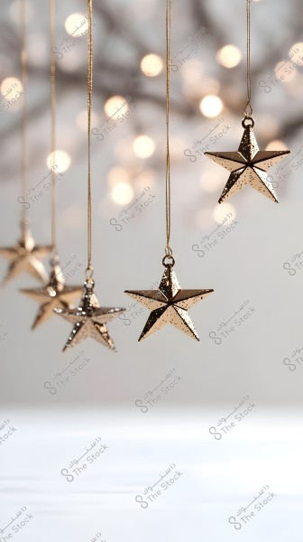 The image features a group of shiny stars made of golden metal, suspended by golden threads. The stars glimmer in the light, with a blurred background focused on soft white lights resembling string lights, giving the image a warm, festive atmosphere.