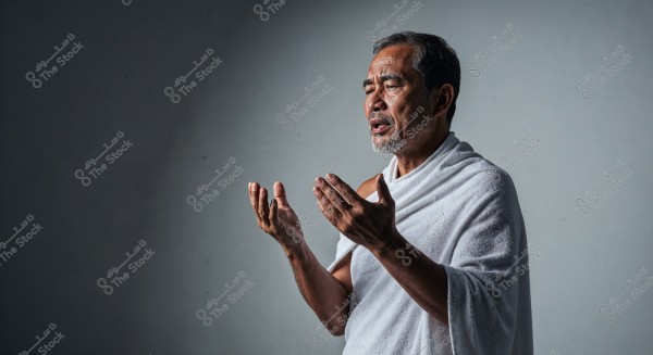 A portrait of an elderly man wearing a white Ihram, indicating participation in religious rituals. The man stands with raised hands as if in prayer, against a gray background. His face reflects an expression of focus and reverence.
