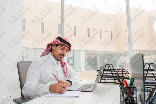 رجل سعودي يرتدي الزي السعودي التقليدي يعمل في مكتب بواجهة زجاجية ويستخدم قلم رصاص لتدوين الملاحظات