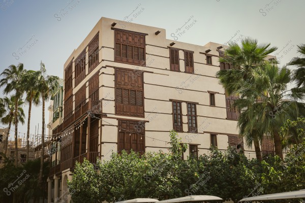 A traditional four-story building in a historic district, featuring authentic Arabic architectural style with ornate wooden windows and balconies. The building is surrounded by palm trees and dense green foliage, reflecting the historic and cultural character of the area under a clear sky.