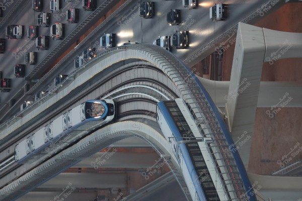 Aerial view showing a modern metro train in blue and white traveling on its winding track, with part of the city traffic in the background where many cars are moving. The image highlights the architectural design of the bridge carrying the train and the adjacent highway.