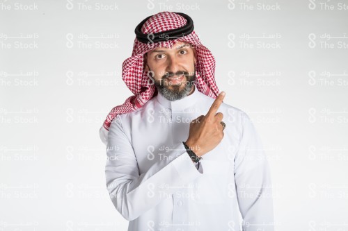 بورتريه رجل سعودي على خلفية بيضاء، يرتدي الثوب وينظر الى الكاميرا مشيرًا بأصبعه على كتفه