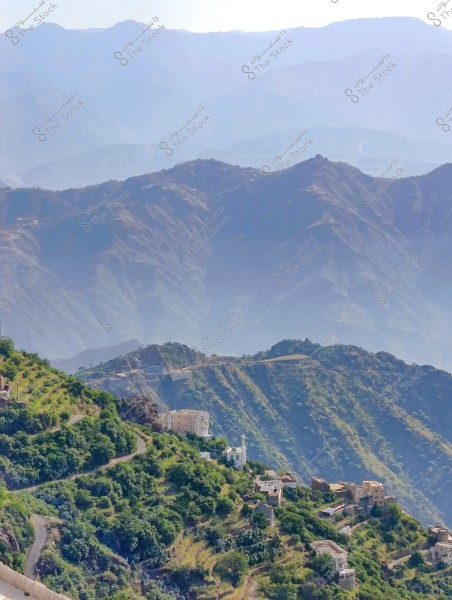 An image showing a scenic view of green mountains in a misty environment. The mountains overlap in the background, with some buildings scattered across the vegetation-covered hills. Roads wind around the contoured lines of the mountains, reflecting the beauty of rural areas in Saudi Arabia.
