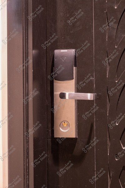 An image of a modern electronic door lock mounted on a dark-colored door. The lock is made of silver metal with a black upper interface featuring a wireless signal icon, a horizontal silver handle, and a key slot at the bottom.