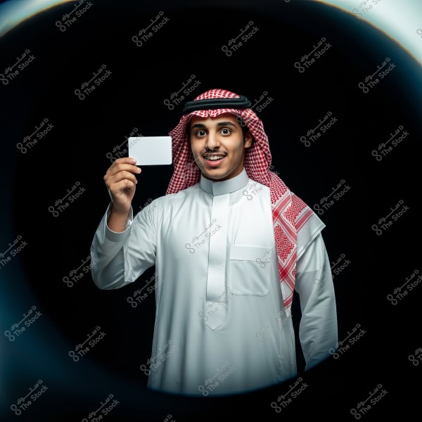A portrait of a man wearing traditional Saudi attire, including a white thobe and a red and white checkered shemagh with a black agal. The man is smiling and holding a white card in his hand, set against a black background.