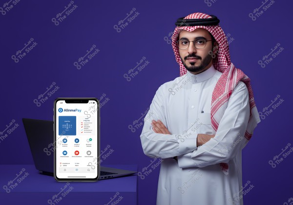 Image of a man wearing traditional Saudi attire with a red and white checkered headscarf and white thobe. He stands with arms crossed against a blue background. Beside him is a smartphone displaying the \"Alinma Pay\" app on its screen, with a laptop in the background.