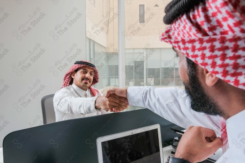 رجلان سعوديان يرتديان الزي السعودي التقليدي يتصافحان أثناء العمل في أروقة المكتب