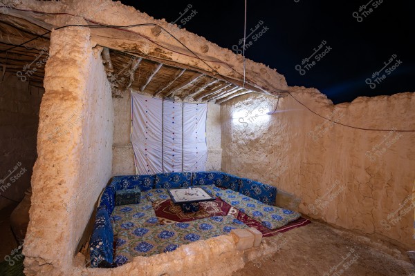 The image shows a traditional open-air seating area of an old mud house, with a gravel floor and a ceiling made of sticks and wood covered with mats. The walls are made of mud bricks, and in the corner, there is traditional Arabic seating adorned with blue cushions and others with vibrant colors. The ground has decorative rugs. A hanging electric lamp illuminates the area.