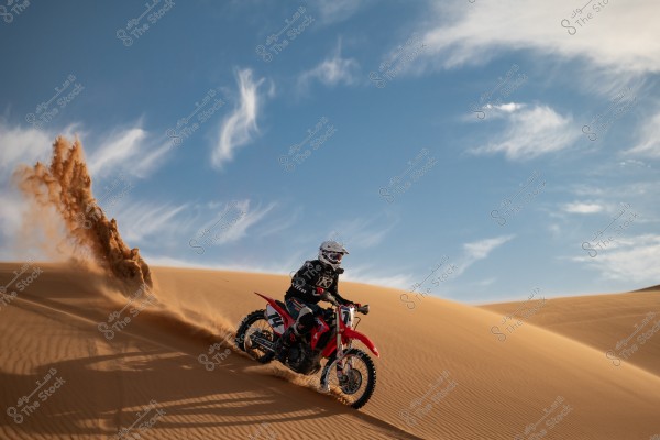 A motorcycle racer wearing protective gear and a white helmet rides a red motorcycle over sand dunes in a desert. Sand sprays into the air as the rider speeds down the dunes. The sky is clear with a few white clouds.