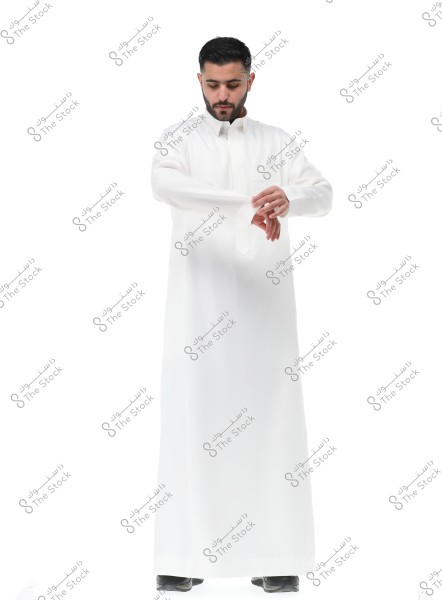 A portrait of a man wearing a traditional white thobe, standing against a white background. He is adjusting the sleeve of the thobe with his hand. The man has a short haircut and a neatly trimmed beard. The attire suggests traditional Arab clothing, possibly from Saudi Arabia.