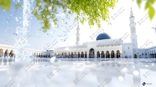 An image of Al-Masjid an-Nabawi in Medina, Saudi Arabia, showcasing the iconic green dome and elegant white minarets. In the foreground, water jets create falling droplets shimmering in the light. At the top of the image, green foliage provides shade over the white surface of the mosque.