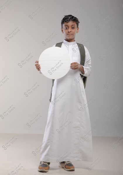 The image shows a child wearing a white thobe and sneakers, carrying a gray backpack. The child is standing in front of a white background holding a round white board. The thobe suggests he might be from the Gulf region, such as Saudi Arabia.