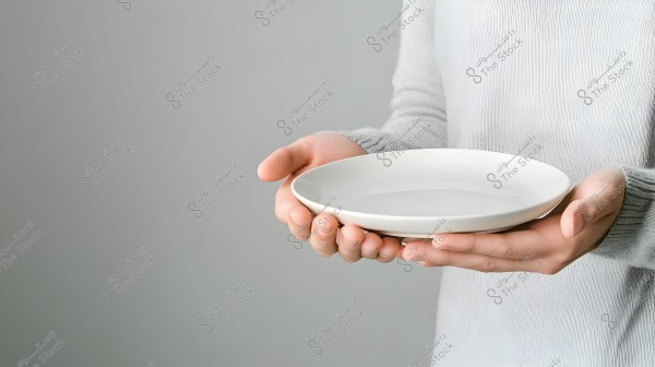 A person wearing a light-colored shirt holding an empty white plate with both hands. The background is plain and lacks detail, drawing focus to the hands and the plate.