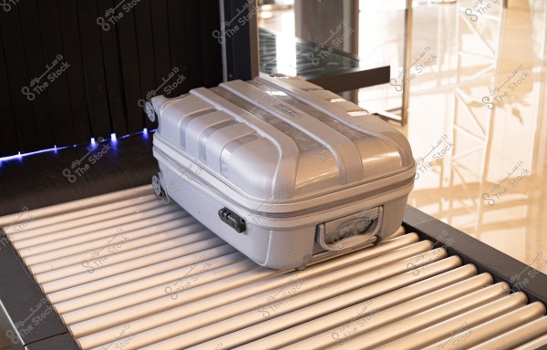 حقيبة سفر رمادية موضوعة على حزام نقل في مطار أو محطة قطار. الحزام مصنوع من بكرات معدنية, ويظهر في الخلفية عاكسًا للضوء مع بعض العناصر المعمارية الحديثة.
