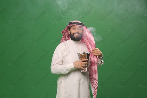 رجل سعودي يقف على خلفية خضراء يرتدي الثوب و الشماغ، يقوم بعمل حركات بيديه ، يبخر نفسه بالطيب و العود، احتفالات العيد