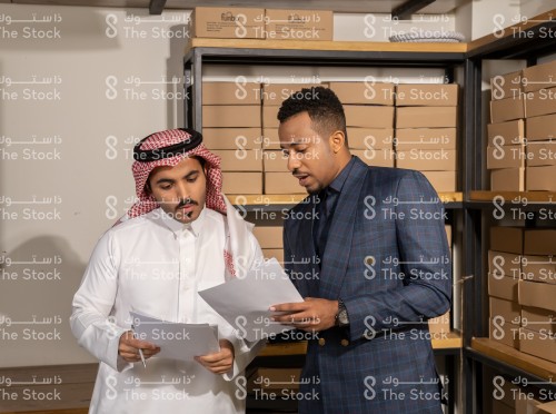 شاب سعودي يرتدي الزي السعودي التقليدي يقف في المستودع مع رجل يرتدي بدلة رسمية بينما يحملان الأوراق وهما يتناقشان وتظهر من خلفهم الرفوف ممتلئة بالصناديق