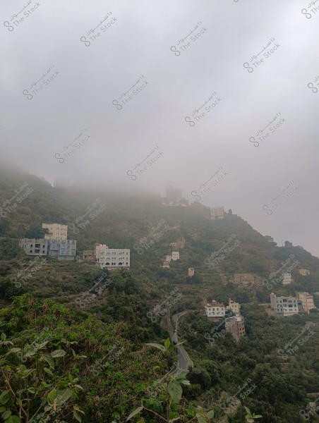 منظر طبيعي لجبل محافظة فيفاء بمنطقة جازان جنوب المملكة العربية السعودية مغطى بالضباب مع مجموعة من البيوت الموزعة على المنحدرات الخضراء. الطرق تتعرج بين المنازل, مما يضيف تفاصيل هندسية إلى المشهد الطبيعي. السماء ملبدة بالغيوم الكثيفة, مما يضفي جوًا غامضًا وساحرًا على الصورة.
