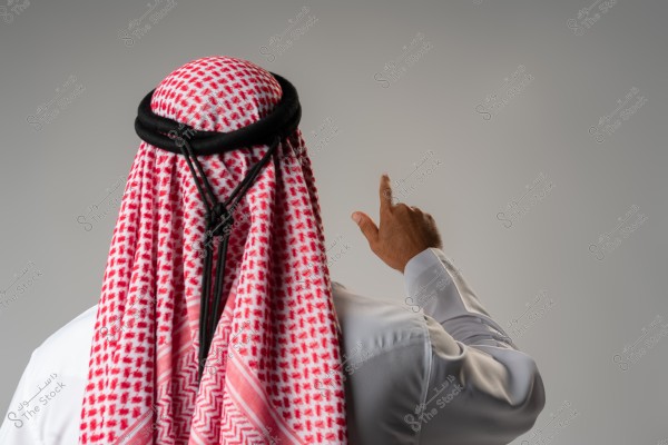 The image shows a man from behind wearing traditional Gulf attire, consisting of a red and white checked keffiyeh and a black agal. The man is pointing upwards with his finger. The background is gray and neutral.