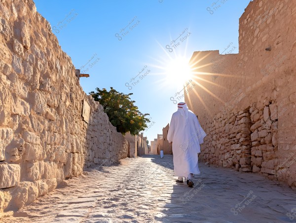 A man wearing traditional Gulf attire, consisting of a white thobe and a red ghutra, walks along a narrow pathway between stone walls in a traditional village under a bright sun. Sun rays shine strongly from the blue sky, and there is a green tree visible on one of the walls.