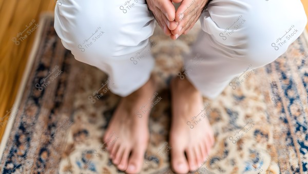 An image showing a person seated on a decorative rug, wearing white trousers, with hands clasped in front and bare feet. The wooden flooring is partially visible on the left side of the image.