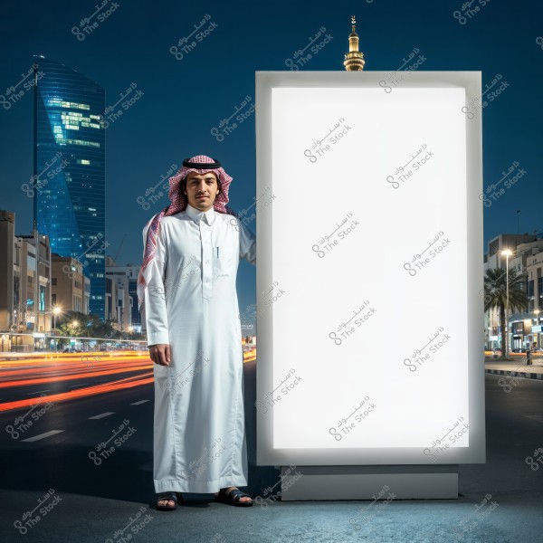 An image of a man standing next to a lit white billboard in an urban street at night. The man is dressed in traditional Saudi attire, including a thobe, ghutra, and agal. Modern buildings and a street with car lights appear in the background, with a prominent tower and a minaret in the skyline.