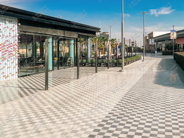 An image of an outdoor area at a modern shopping center. The space features a black and white checkerboard-patterned tiled floor, lined with closed umbrellas and seating arrangements on one side. It is surrounded by palm trees and neatly trimmed ornamental plants. There are also glass walls of a building on the left with white and pink decorations. The sky is clear with a few scattered clouds.