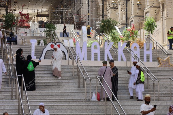 A group of people walking on a large staircase in Mecca. In the background, there is a large sign that says \"I ♥ MAKKAH\" with a heart design. The people are wearing traditional clothing, some in black abayas and others in white thobes. The area is decorated with plants and hanging lights.